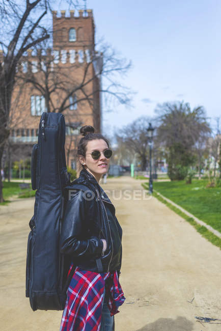 Вид сбоку на молодую улыбающуюся хипстершу, гуляющую в парке в солнечный день с гитарой на спине — стоковое фото