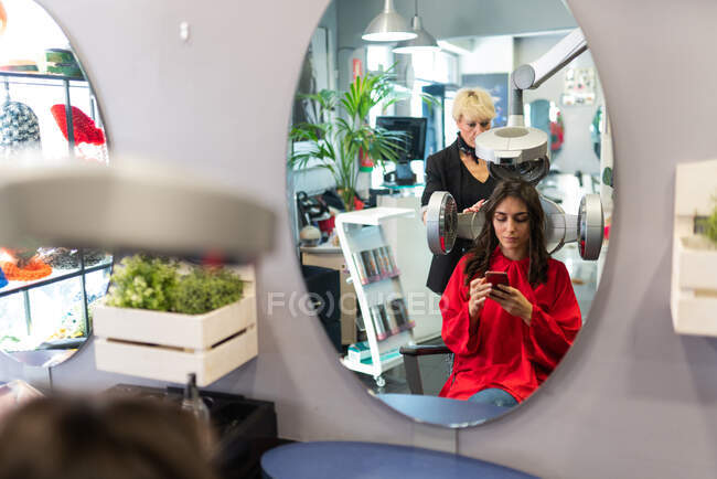 Vista lateral de la alegre dama sosteniendo el teléfono móvil y secando pelos en el salón de peluquería - foto de stock