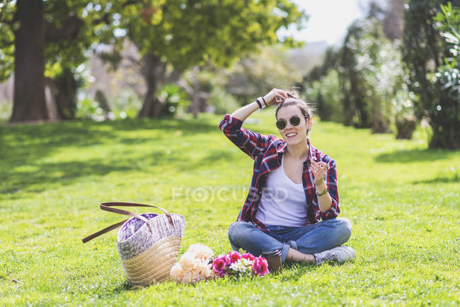 Frontansicht einer jungen Hipsterfrau, die in einem Park auf Gras sitzt, während sie eine Blume in der Hand hält und an einem sonnigen Tag lächelt — Stockfoto