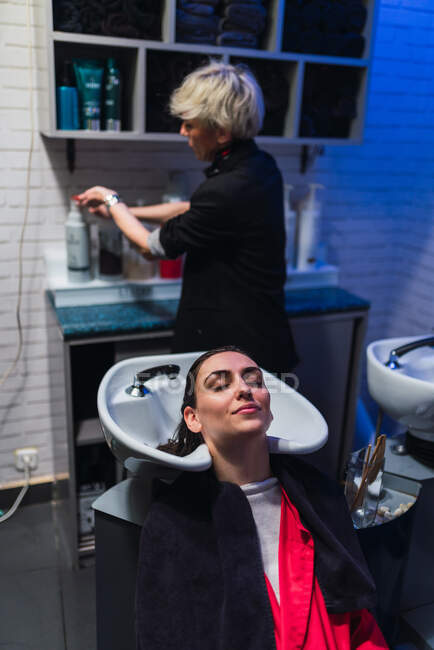 Aged stylist washing hairs of attractive lady near sink in hairdressing salon — Stock Photo