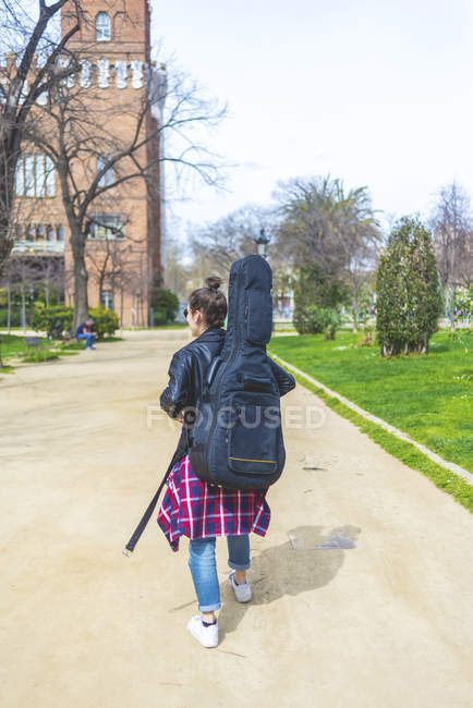 Rear view of a young hipster woman walking in a park in sunny day while carrying a guitar on back — Stock Photo