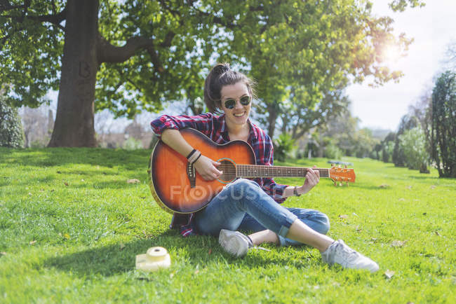 Vista frontal de uma jovem hipster usando óculos de sol, sentada na grama em um parque enquanto aprecia tocar guitarra — Fotografia de Stock