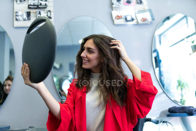 Stylist holding mirror with reflection of attractive cheerful lady in hairdressing salon — Stock Photo