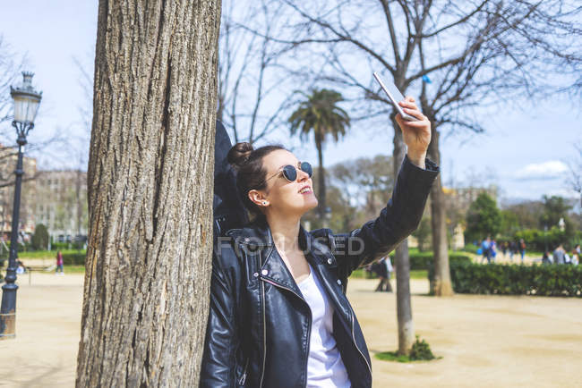 Вид збоку на жінку, що стоїть і спирається на дерево в парку в сонячний день, використовуючи мобільний телефон, щоб взяти селфі — стокове фото