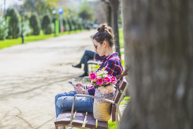 Вид збоку на молоду хіпстерку, яка сидить на лавці в парку, розслабляючись в сонячний день, використовуючи мобільний телефон — стокове фото