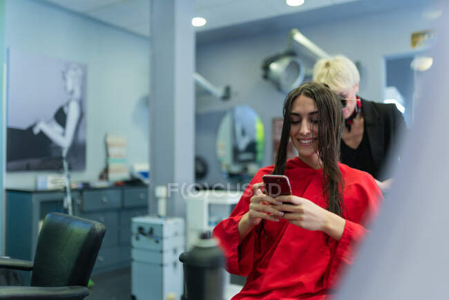 Молодая женщина с мобильным телефоном и сидя на стуле с красивой прической в парикмахерской — стоковое фото