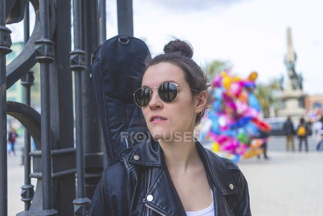 Vista frontal de una joven mujer hipster riendo de pie y apoyada en una valla en el parque en un día soleado mientras mira hacia otro lado - foto de stock