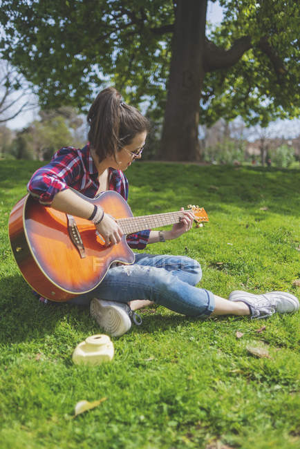 Vista frontal de una joven mujer hipster con gafas de sol, sentado en la hierba en un parque mientras disfruta tocando la guitarra - foto de stock