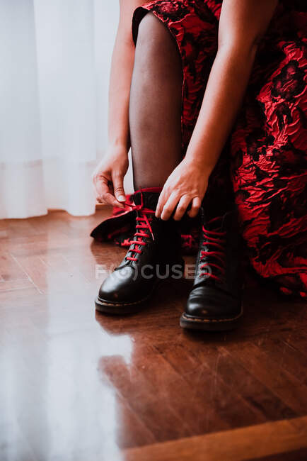 Senhora de colheita em vestido vermelho atacando botas pretas no quarto com piso de madeira perto de cortinas — Fotografia de Stock