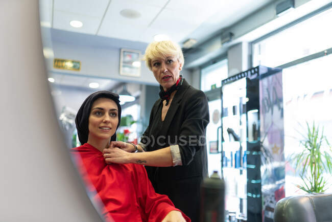 Reflet de styliste âgé debout près de dame attrayante avec serviette sur la tête dans le salon de coiffure — Photo de stock