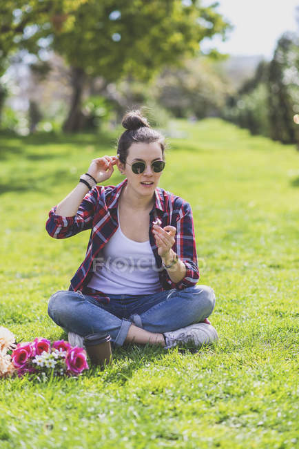 Вид спереду молода хіпстерка, що сидить на траві в парку, тримаючи квітку в сонячний день — стокове фото