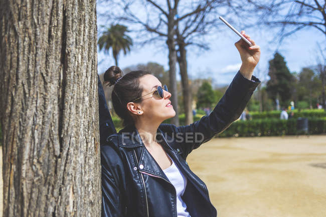 Side view of woman standing and leaning on a tree at park in sunny day while using a mobile phone to take a selfie — Stock Photo