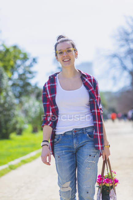 Frontansicht einer jungen lächelnden Hipsterfrau, die an sonnigen Tagen in einem Park spaziert und dabei einen bösen Korb hält — Stockfoto