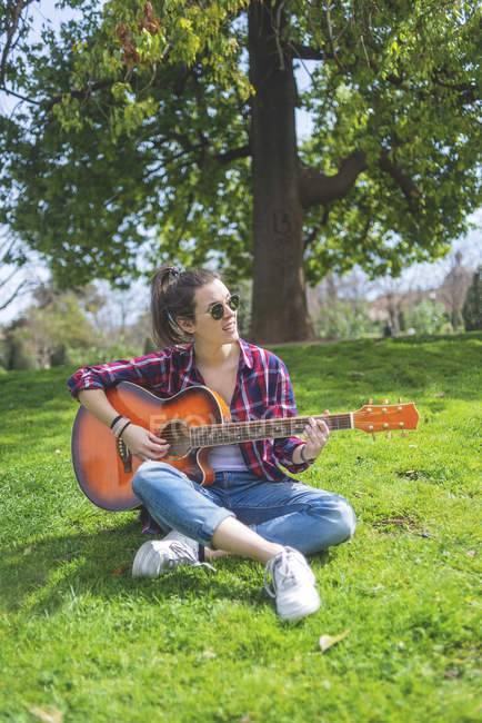 Vista frontal de una joven mujer hipster con gafas de sol, sentado en la hierba en un parque mientras disfruta tocando la guitarra - foto de stock