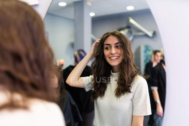 Reflection of young beautiful lady touching hair looking at mirror in hairdressing salon — Stock Photo