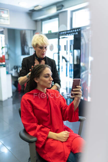 Giovane donna con telefono cellulare e seduta sulla sedia con bella acconciatura nel salone di parrucchiere — Foto stock