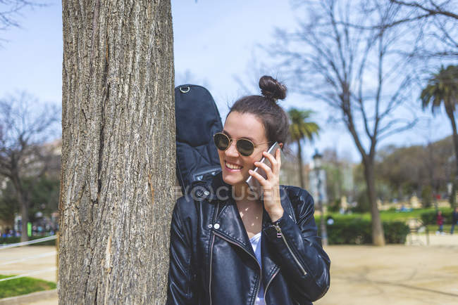 Вид сбоку на молодую смеющуюся хипстершу, стоящую и опирающуюся на дерево в парке в солнечный день, используя мобильный телефон — стоковое фото