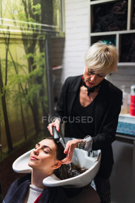 Stilista anziano che lava capelli di signora attraente vicino a lavandino in salone di parrucchiere — Foto stock