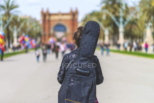 Vue arrière d'une jeune femme hipster marchant dans un parc par temps ensoleillé tout en portant une guitare sur le dos — Photo de stock