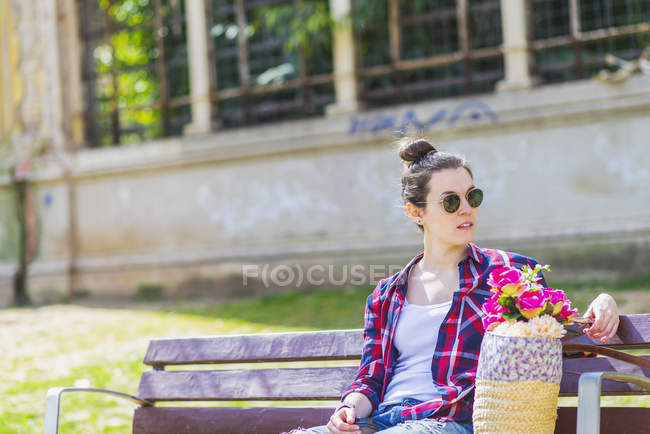 Вид спереду молода хіпстерка, що сидить на лавці в парку, розслабляється в сонячний день, дивлячись далеко — стокове фото