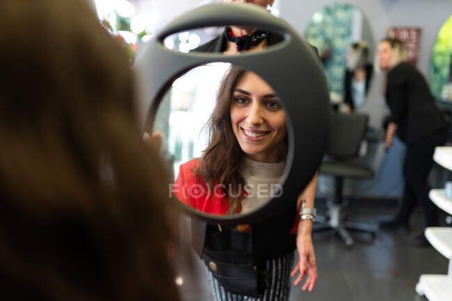 Stylist hält Spiegel mit Reflexion der attraktiven, fröhlichen Dame im Friseursalon — Stockfoto