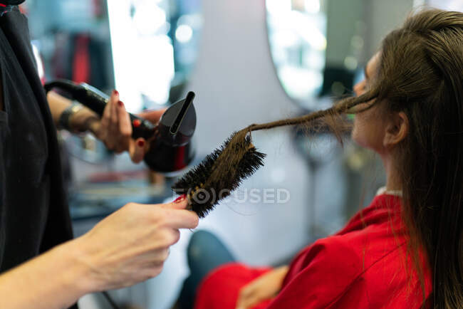 Crop estilista usando secador e pente e fazer penteado para mulher atraente no salão de cabeleireiro — Fotografia de Stock