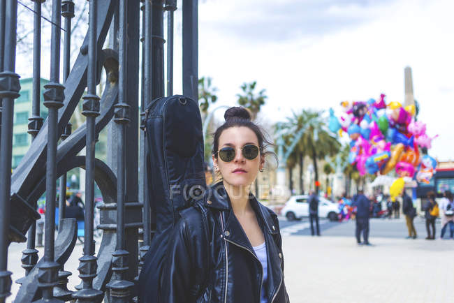 Vista frontale di una giovane donna hipster ridente in piedi e appoggiata su una recinzione nel parco durante la giornata di sole mentre guarda altrove — Foto stock