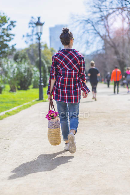 Vista trasera de una joven hipster caminando en un parque en un día soleado mientras sostiene una cesta malvada - foto de stock
