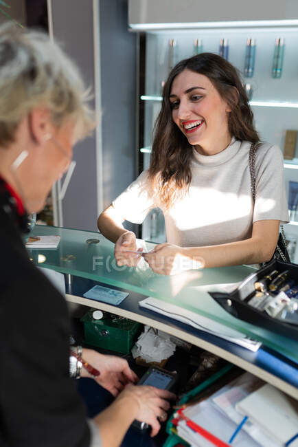 Side view of happy lady with makeup paying by plastic card near cheerful stylist in hairdressing salon — Stock Photo