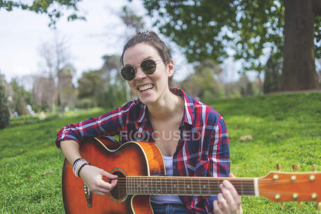 Vista frontale di una giovane hipster che indossa occhiali da sole, seduta sull'erba in un parco mentre si diverte a suonare la chitarra — Foto stock