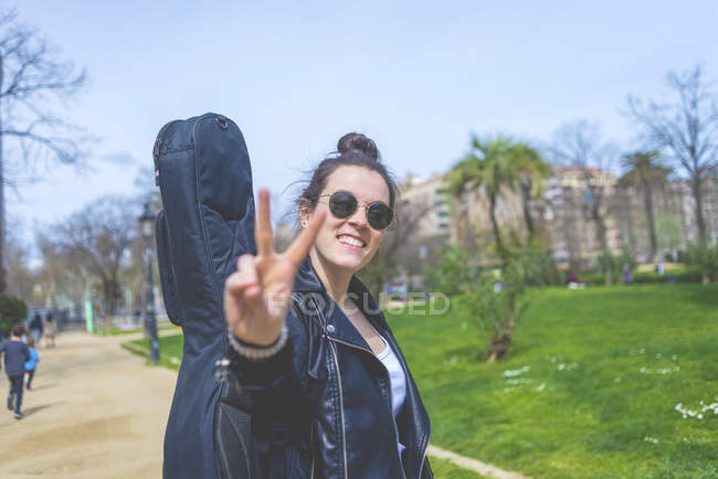 Vista lateral de la mujer caminando en un parque en un día soleado mientras lleva una guitarra en la espalda y un gesto signo de victoria - foto de stock
