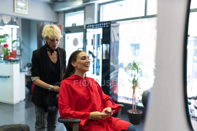 Jovem mulher com telefone celular e sentada na cadeira com belo penteado no salão de cabeleireiro — Fotografia de Stock