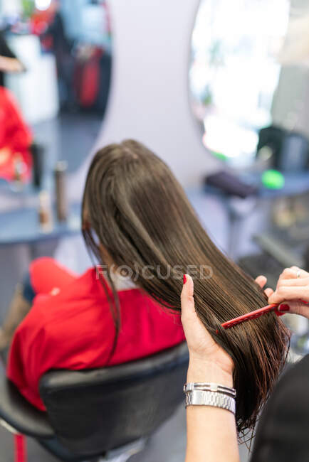 Crop hands of stylist combing hair of brunette woman sitting on chairs in hairdressing salon — Stock Photo