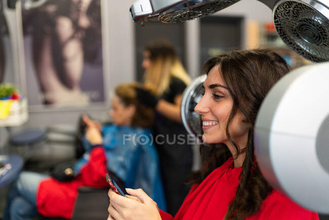 Вид збоку весела дама тримає мобільний телефон і сушильні волоски в перукарні — стокове фото
