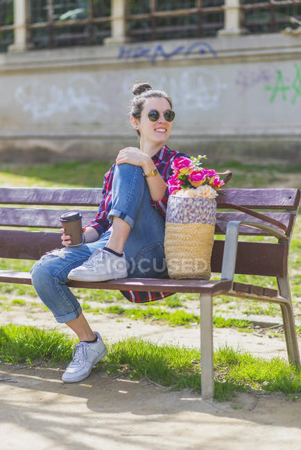 Вид спереди на молодую хипстершу, сидящую на скамейке в парке и отдыхающую в солнечный день. — стоковое фото