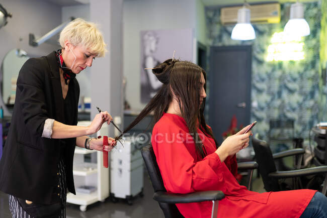 Молода жінка з мобільним телефоном і сидить на стільці з красивою зачіскою в перукарні салону — стокове фото