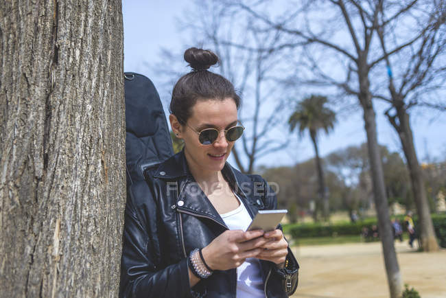 Vista lateral de la mujer de pie y apoyado en un árbol en el parque en el día soleado mientras se utiliza un teléfono móvil - foto de stock