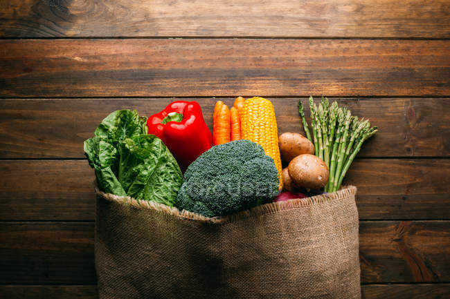 Surtido de verduras frescas crudas con saco en mesa de cocina de madera - foto de stock