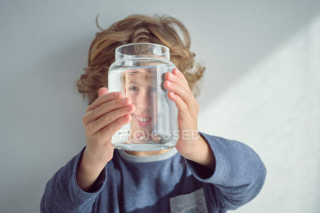 Menino bonito sorrindo e segurando frasco de vidro com água limpa na frente do rosto, enquanto está contra a parede branca — Fotografia de Stock