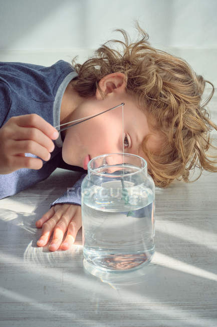 Petit garçon joyeux trempant minuscule canne à pêche dans un bocal en verre avec de l'eau propre tout en étant couché sur le sol — Photo de stock