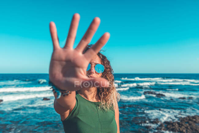 Dame à la mode dans les lunettes de soleil montrant geste d'arrêt à la caméra tout en se tenant près de la belle mer ondulante contre ciel bleu sans nuages sur une journée ensoleillée — Photo de stock
