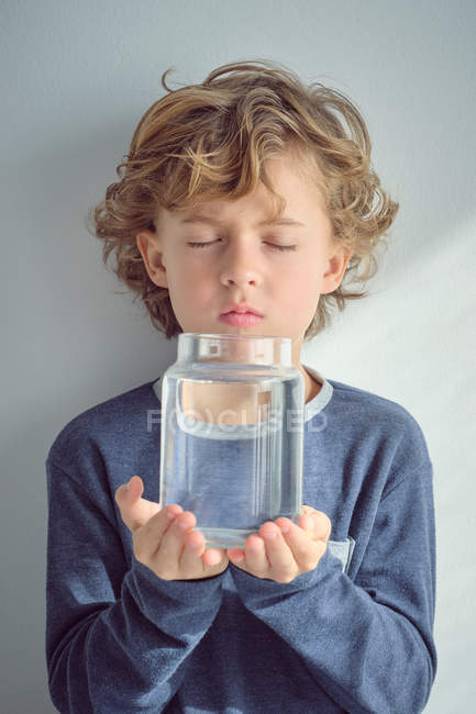 Маленький мальчик с закрытыми глазами держит стеклянную вазу с прозрачной водой, стоя напротив белой стены — стоковое фото