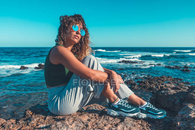 Side view of attractive dreamy female in trendy outfit and sunglasses embracing knees and looking away while sitting on shore near waving sea — Stock Photo