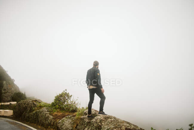 Back view of male in casual outfit standing on rocky cliff near countryside road on foggy day in Spain — Stock Photo