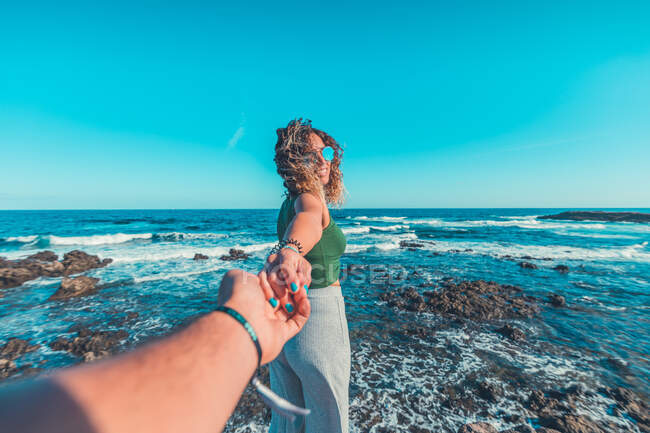 Pretty young female smiling and holding crop hand of anonymous friend while standing against majestic waving sea and clear blue sky — Stock Photo