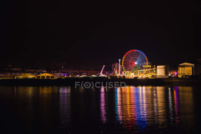 Glowing amusement park with big observe wheel in colorful lights reflecting in water of city channel in night — Stock Photo