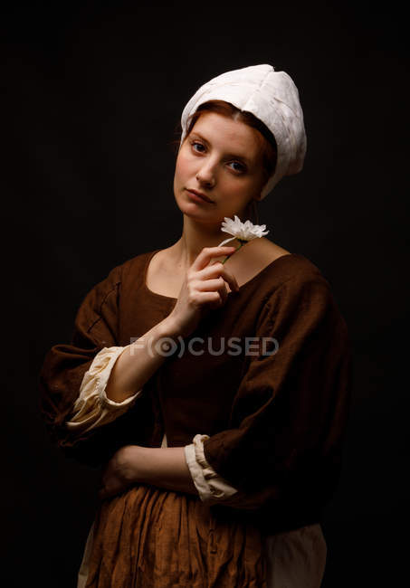Attractive woman in medieval dress holding small flower and looking in camera on black background. — Stock Photo
