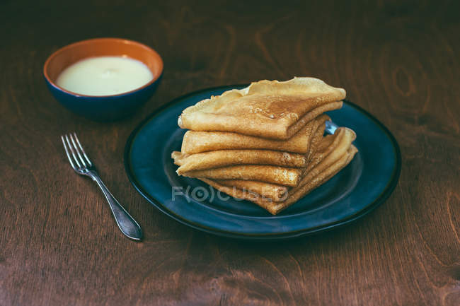 Plate with pile of delicious blini and bowl of sour cream on timber tabletop. — Stock Photo