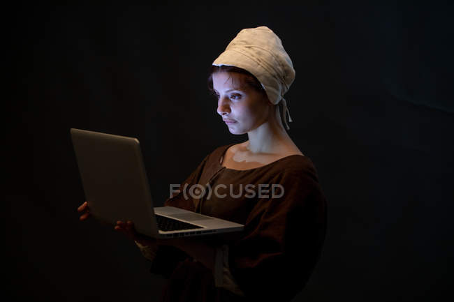 Side view of attractive female in simple medieval dress and bonnet browsing modern laptop. — Stock Photo