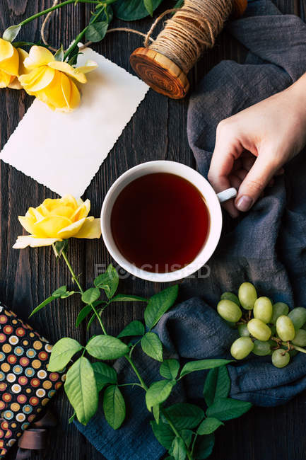 From above hand of female holding cup of aromatic tea near elegant yellow roses and empty note over wooden tabletop. — Stock Photo